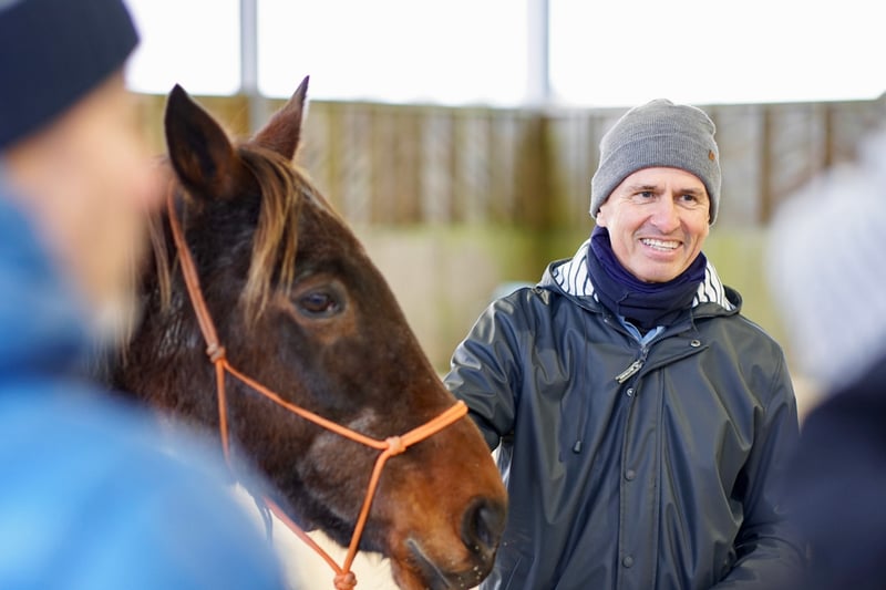 Smiling man and a horse in a barn. The man is interacting with two other people that are out of focus and we can't see - but he looks engaged and happy.